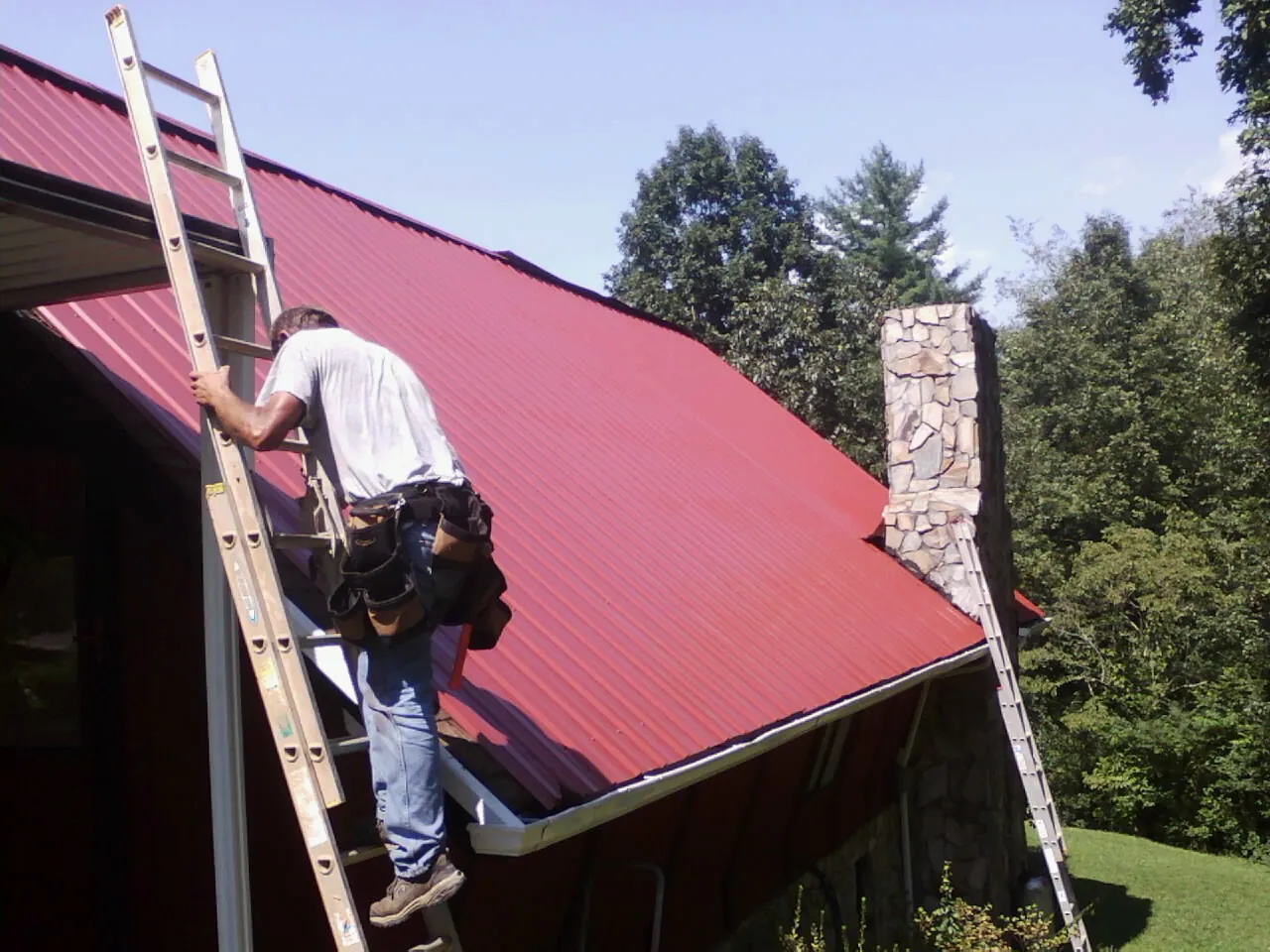 Roofer on a ladder beside a red metal roof and stone chimney — Soffit & Fascia Repair by Roofing Brothers in Kimmell
